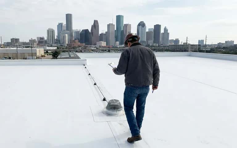 Inspector with a clipboard walking a white membrane roof beside a roof drain