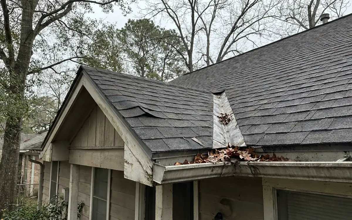 Weathered rental roof edge with a lifted shingle and leaves in the gutter