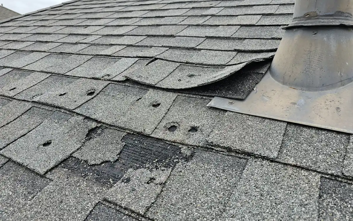 Roof shingles with scattered impact marks, lifted edges, and vent flashing after a storm
